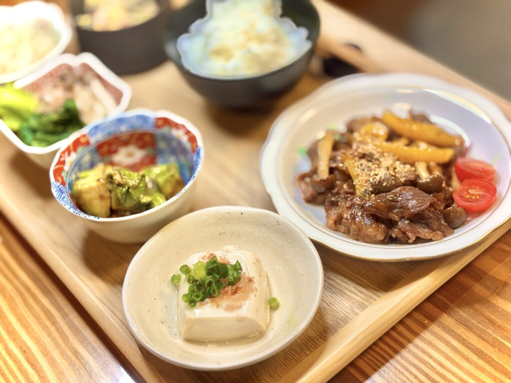 木のトレーに並んだ夕食セット。中央には冷奴、その奥にはアボカドの和え物や小松菜のおひたし。右側には牛肉ときのこのプルコギが盛られ、黄色パプリカとプチトマトが彩りを添えている。和風小鉢とご飯が揃った、居酒屋風の献立。