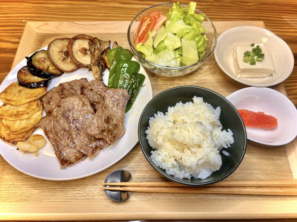木のトレーに並んだ焼肉定食。メインは牛肉と野菜のソテーで、付け合わせにしいたけやピーマン、ポテト。小鉢には冷奴とからし明太子、さらにサラダと白ご飯が揃い、バランスよく盛り付けられた食卓。