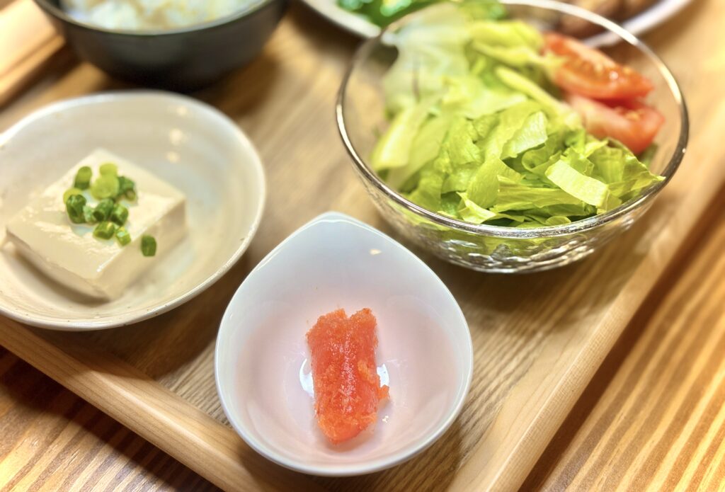 小鉢に盛られた副菜のセット。ガラスの器にはレタスとトマトのサラダ、白い器には刻みねぎをのせた冷奴、しずく型の器にはからし明太子が盛られている。シンプルながら彩り豊かな副菜が並んだ食卓。