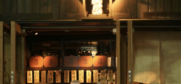 木の温もりある外観の居酒屋入口。暖簾の奥には提灯の明かりと手書きの品書き札が並び、落ち着いた雰囲気の店内がのぞける。