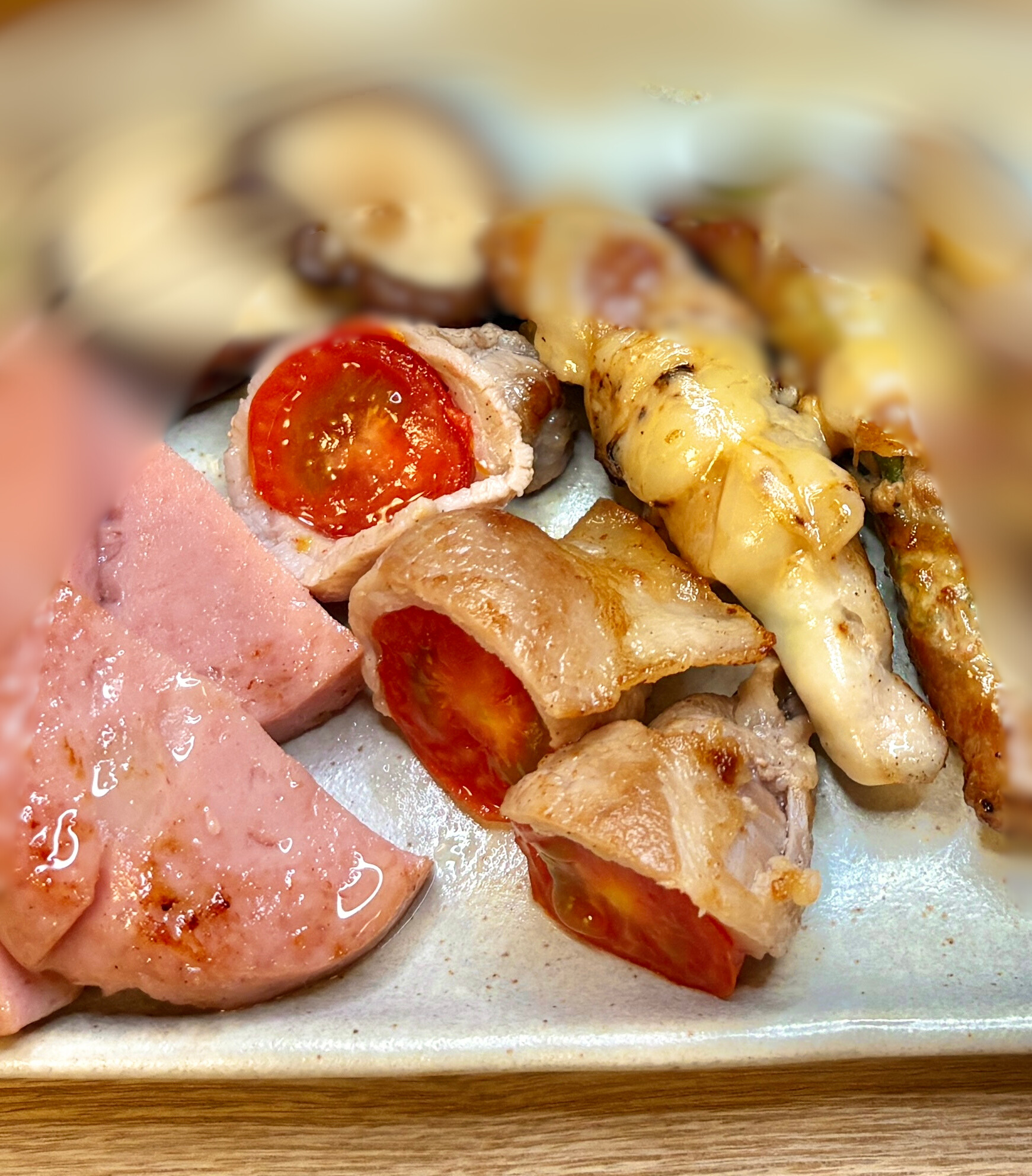 ミニトマトの豚肉巻きが中央にくっきり映り、周囲のボロニアソーセージやしいたけ、チーズ巻きが柔らかくぼけた構図。温かみのある照明で美味しさが引き立つ一枚