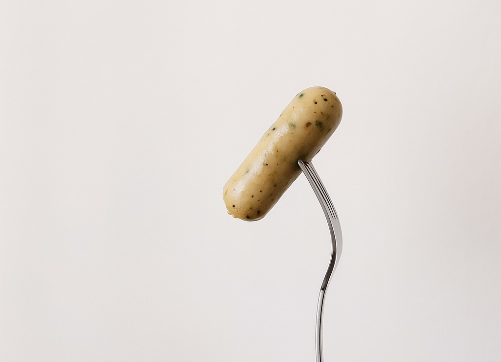 フォークに刺したゆかり入りソーセージをシンプルな背景の中で撮影。 ソーセージの表面にはゆかりの粒が見え、滑らかな質感とナチュラルな照明が際立つミニマルな構図。