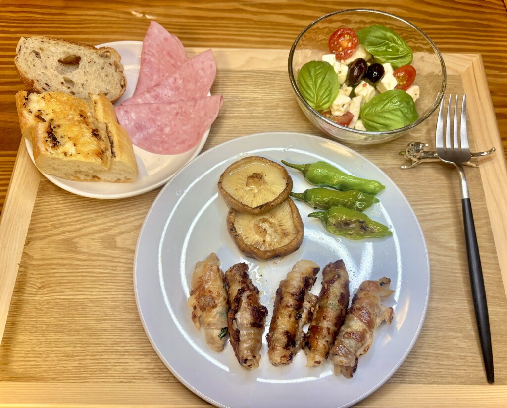 木製トレーに並んだ夕食プレート。豚肉の紫蘇巻き、椎茸とししとうのソテー、ブラックオリーブとチーズとトマトのバジルサラダ、ビアソーセージハム、ビゴのフランスパンが彩りよく盛り付けられている