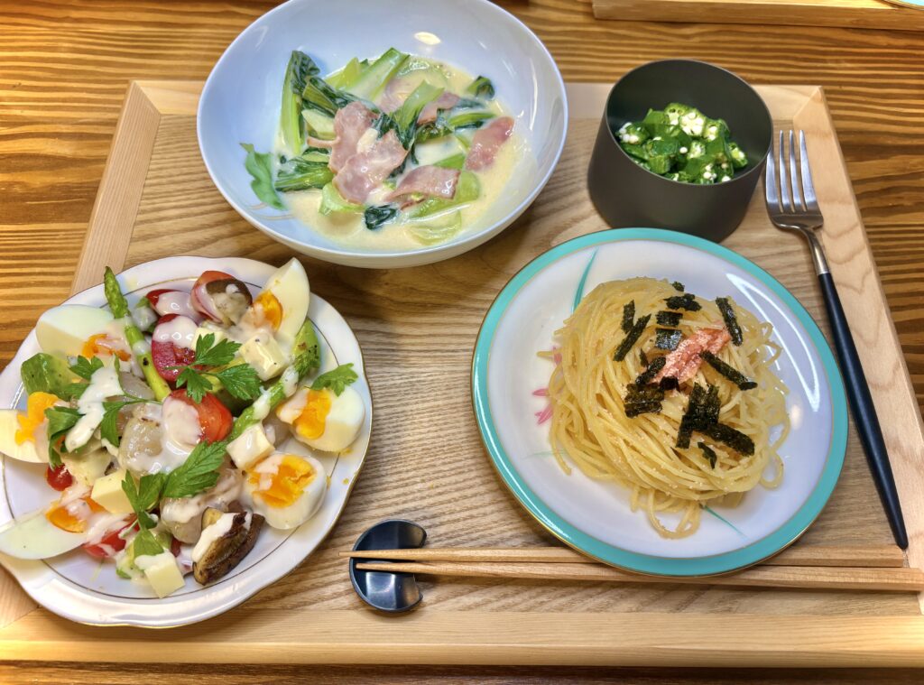 木のトレーに並べられた晩ごはん。チンゲンサイとベーコンのクリーム煮、明太パスタ、ゆで卵やアスパラ入りの彩りサラダ、小鉢のオクラ和えが添えられている。