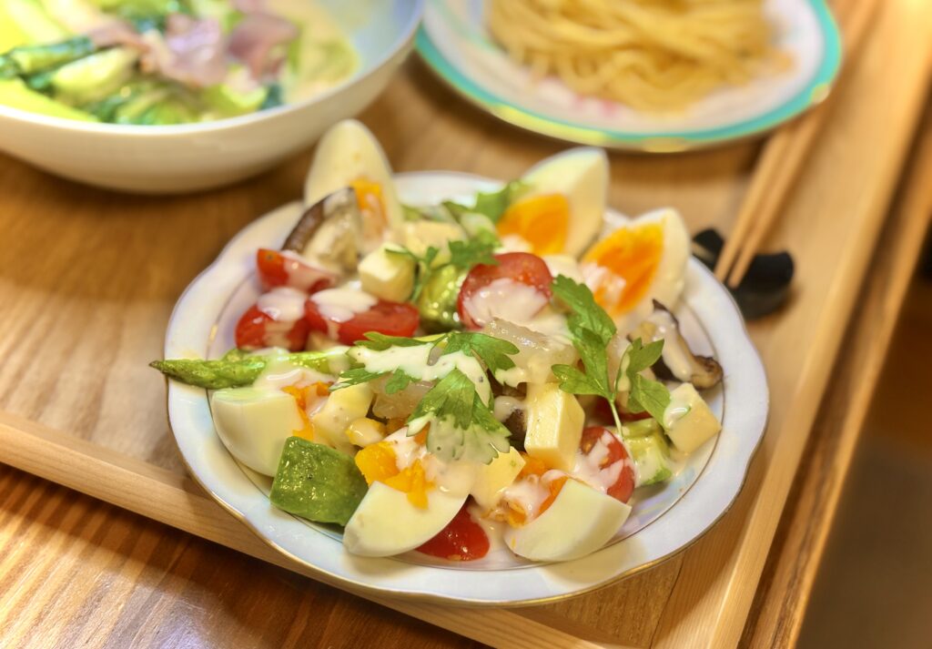 白い皿に盛られた彩りサラダ。ゆで卵、アスパラ、プチトマト、チーズ、しいたけ、葉野菜にドレッシングがかかっている。奥にはチンゲンサイのクリーム煮と明太パスタも見える。
