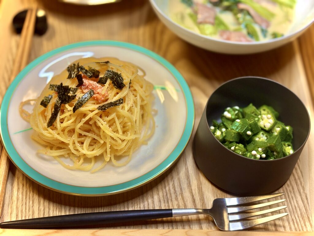 丸皿に盛られた明太パスタに刻み海苔がのっている。隣には黒い器に入った刻みオクラの和え物。背景にはチンゲンサイのクリーム煮やサラダも見える。