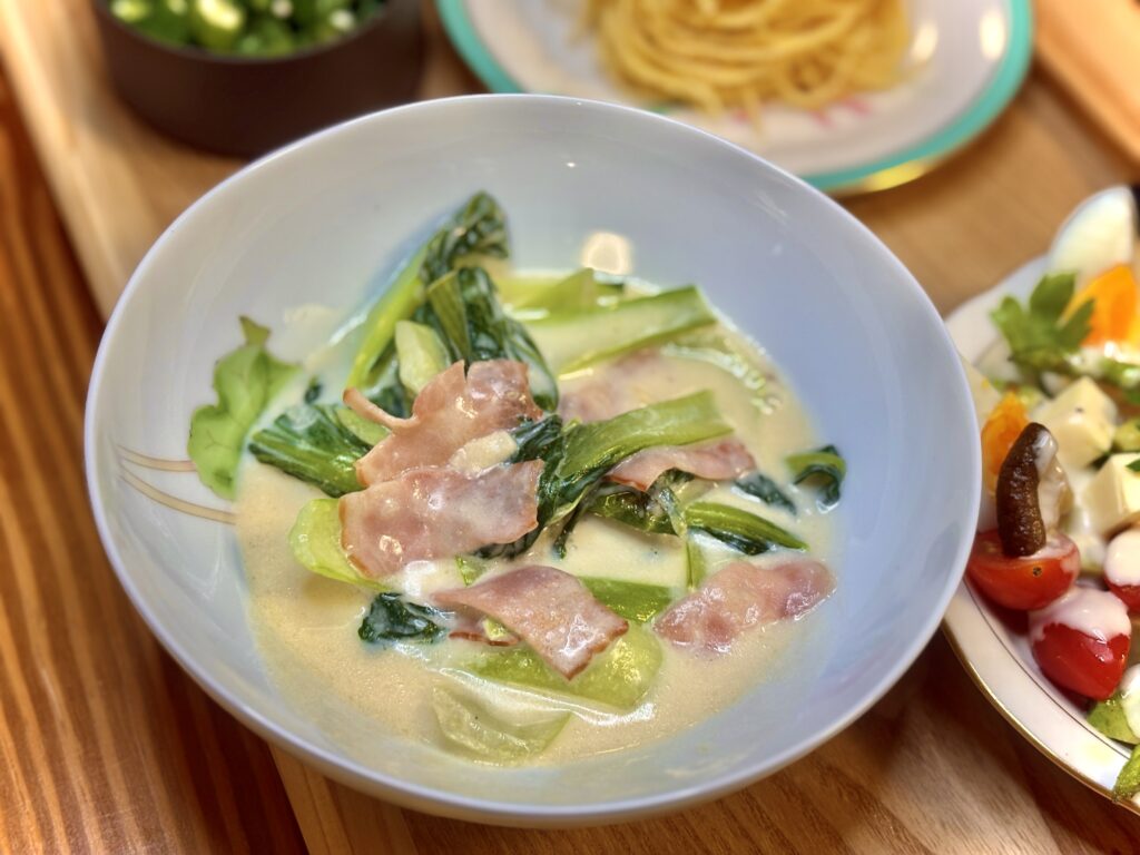 白い器に盛られたチンゲンサイとベーコンのクリーム煮。ミルキーなソースが具材を包み、彩り野菜のサラダや明太パスタと一緒に並べられている。