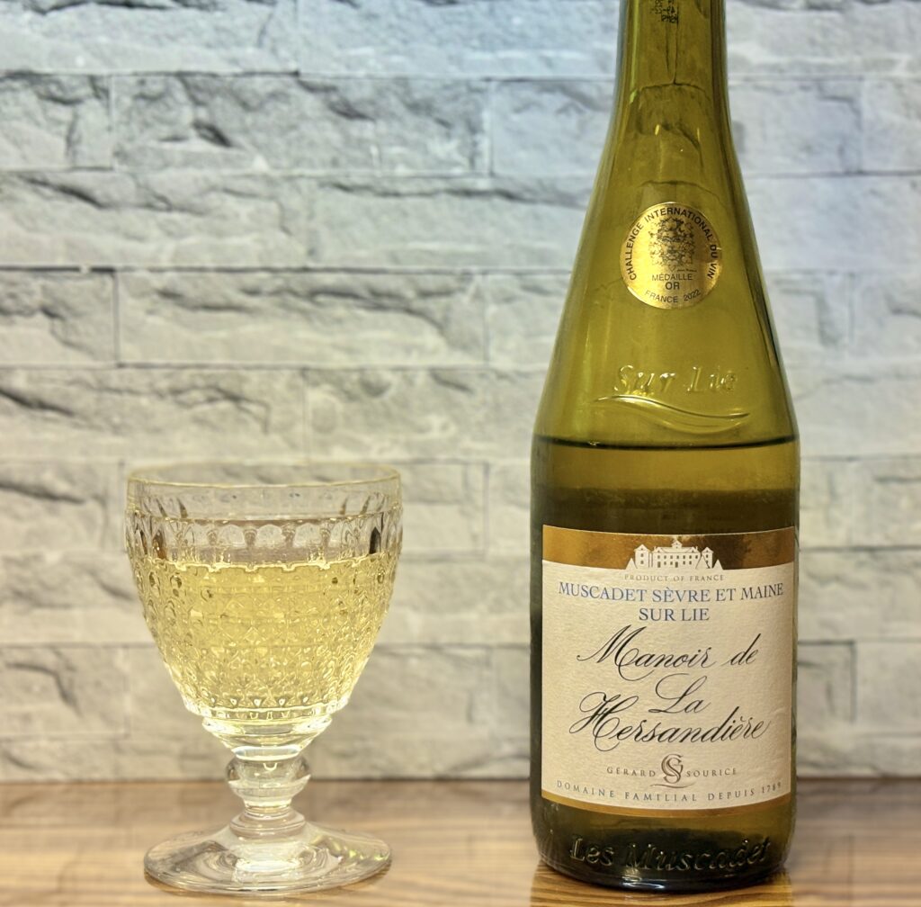 フランス産白ワイン『Muscadet Sèvre et Maine Sur Lie』のボトルと、注がれた白ワインのグラスが木のテーブルに置かれている。背景はグレーの石壁風。