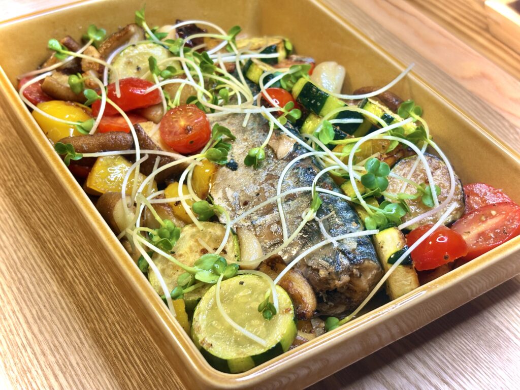 オーブン皿に盛りつけられた鯖のアヒージョ。ズッキーニ、ミニトマト、パプリカ、きのこなどの彩り野菜がたっぷり入り、上にはかいわれが散らされている。オリーブオイルの艶が食欲をそそる一品。