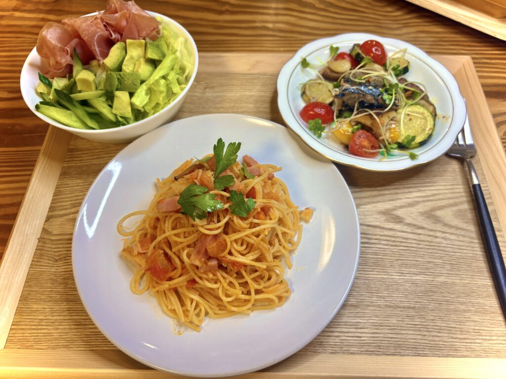 木製トレーに並べられた洋風ディナー。中央にはトマトソースのスパゲッティ、左には生ハムとアボカド、レタス、きゅうりのサラダ、右にはサバと彩り野菜のアヒージョ風小鉢が並び、色鮮やかで食欲をそそる盛り付け。