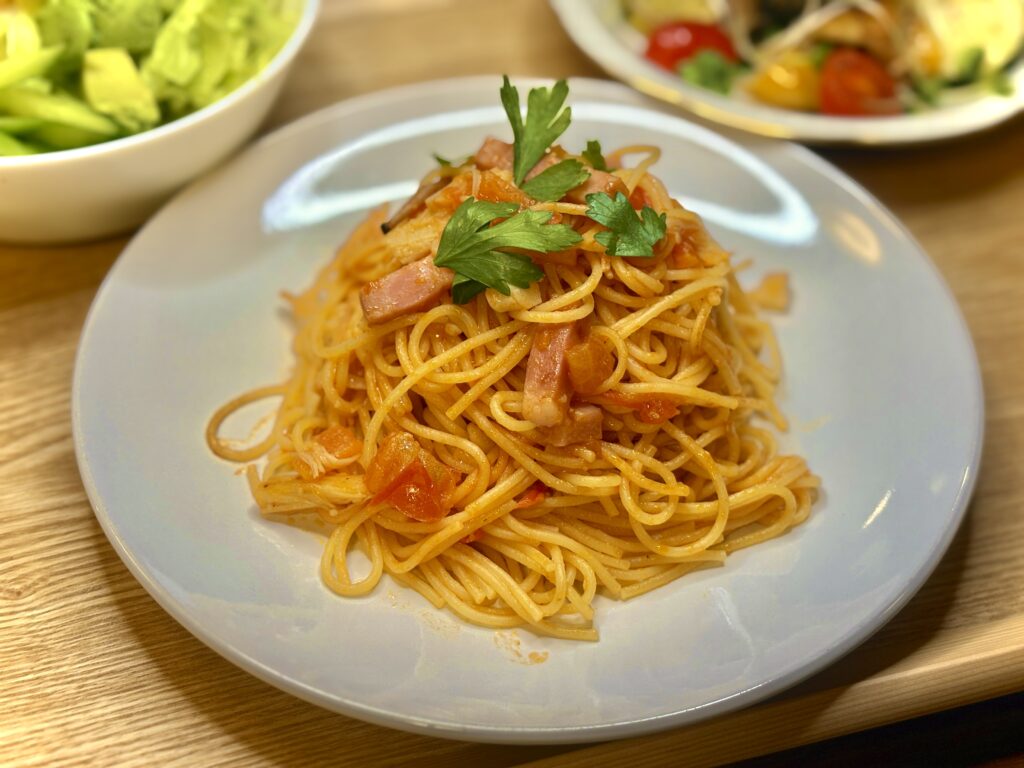 白いお皿に盛られたトマトソースのスパゲッティ。ベーコンと玉ねぎ、トマトが絡み、上にはパセリが添えられている。奥にはアボカドサラダとカラフルな副菜が見える。