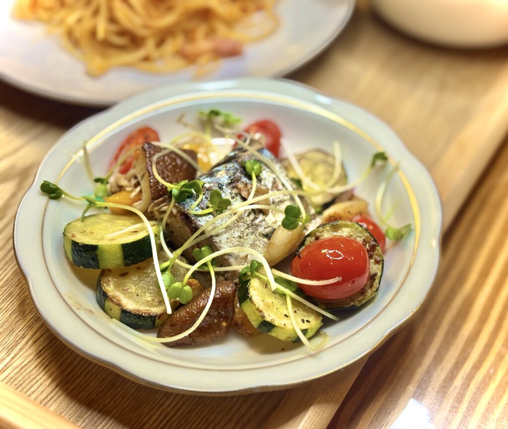 小皿に盛られた鯖と彩り野菜のアヒージョ。ズッキーニやトマト、きのこがカラフルに並び、かいわれの緑がアクセントになっている。奥にはトマトパスタがぼんやり見え、食卓の温かい雰囲気が伝わる。