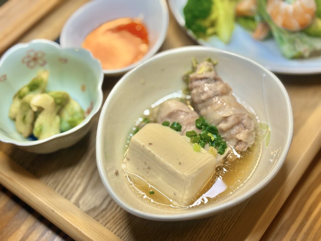柚豆腐と白菜の豚肉巻きを出汁で煮た料理。優しい色合いの器に盛られ、ねぎが添えられている。隣には枝豆の天ぷらとスイートチリソースの小鉢も並び、落ち着いた木のトレイの上に和風アジアンな食卓が広がっている。