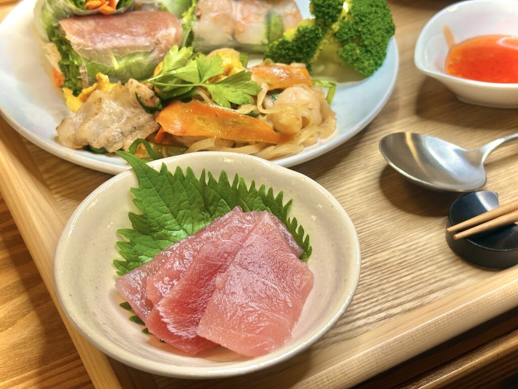 大葉を敷いた皿に盛り付けられたまぐろの刺身。奥には、エビと生ハムの生春巻き、パッタイ、ブロッコリー、スイートチリソースの小皿が並び、木のトレイの上に彩り豊かなアジアンディナーが広がっている。