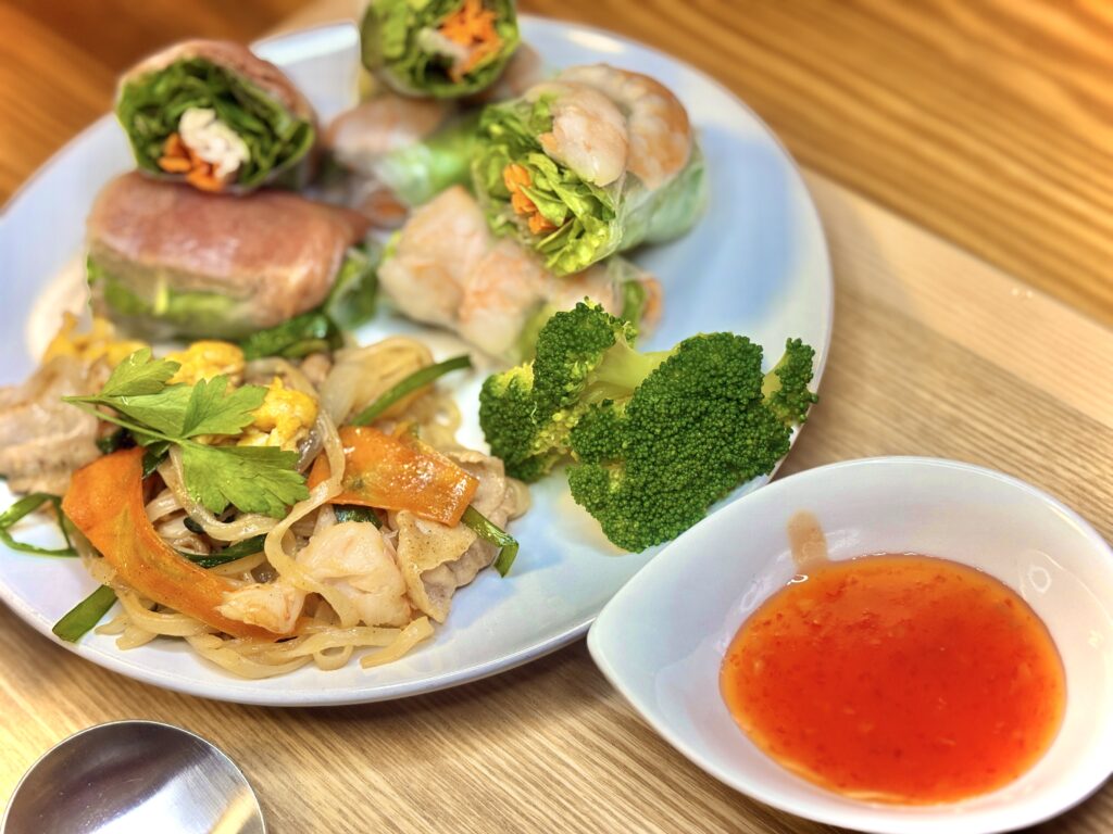 エビと鶏むね肉・アボカドの生春巻き、チーズ生ハムの生春巻き、野菜入りパッタイ、ブロッコリーを盛り合わせたプレート。横にはスイートチリソースが添えられ、カフェ風の彩り豊かなアジアンディナー。