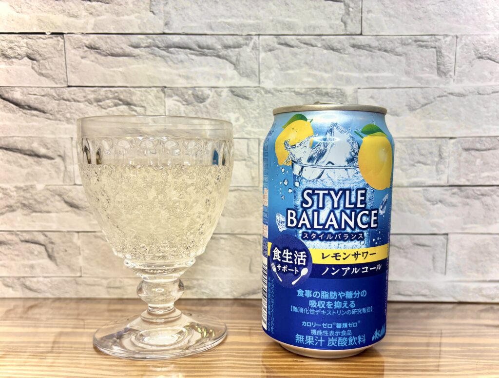 アサヒのノンアルコール飲料「STYLE BALANCE〈レモンサワー〉」の缶と、炭酸の気泡が美しく立ち上るグラス。明るい木のテーブルと白いレンガ調の背景に映える、爽やかなレモンイエローとブルーのパッケージが印象的。