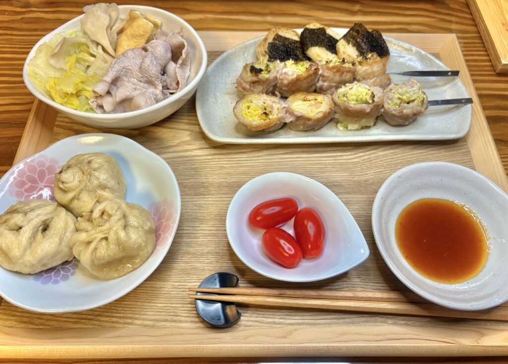 木のトレーに並べられた晩ごはんの献立。左手前にふっくらとした老詳記の豚まん3個、奥には白菜と豚バラの冷しゃぶ、右奥には白菜の豚バラ巻き串と長芋の磯部焼き、中央にミニトマト、右下にはタレ皿。家庭の温かさが伝わる、彩り豊かな和風の夕食。