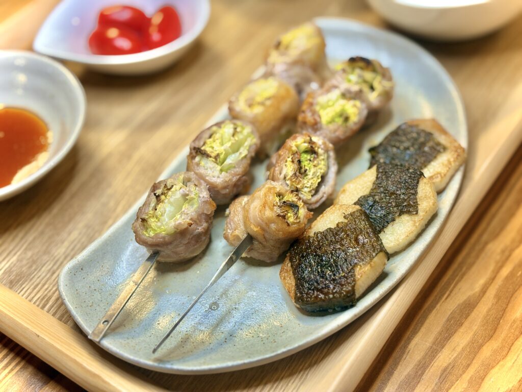 長方形の皿に並んだおかず。手前は磯の香りが広がる長芋の磯部焼き、奥には白菜を豚バラで巻いた串焼き。断面には野菜の彩りが見え、照り焼き風の焼き目が香ばしい。奥にはタレ皿とミニトマトが添えられた温かな食卓の一皿。