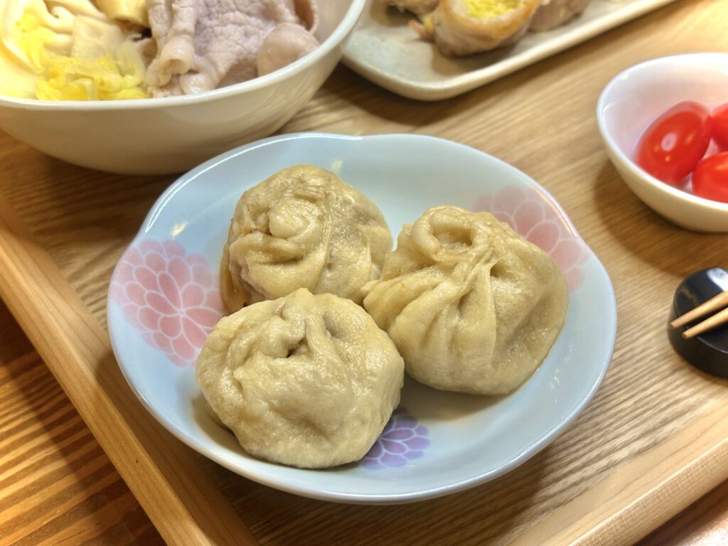花柄の小皿に並べられた老詳記の豚まん3個。ふっくらとした生地から肉汁がにじむような質感で、湯気の立つ温かさが伝わる。奥には冷しゃぶやトマトなど、晩ごはんの副菜がちらりと見える。