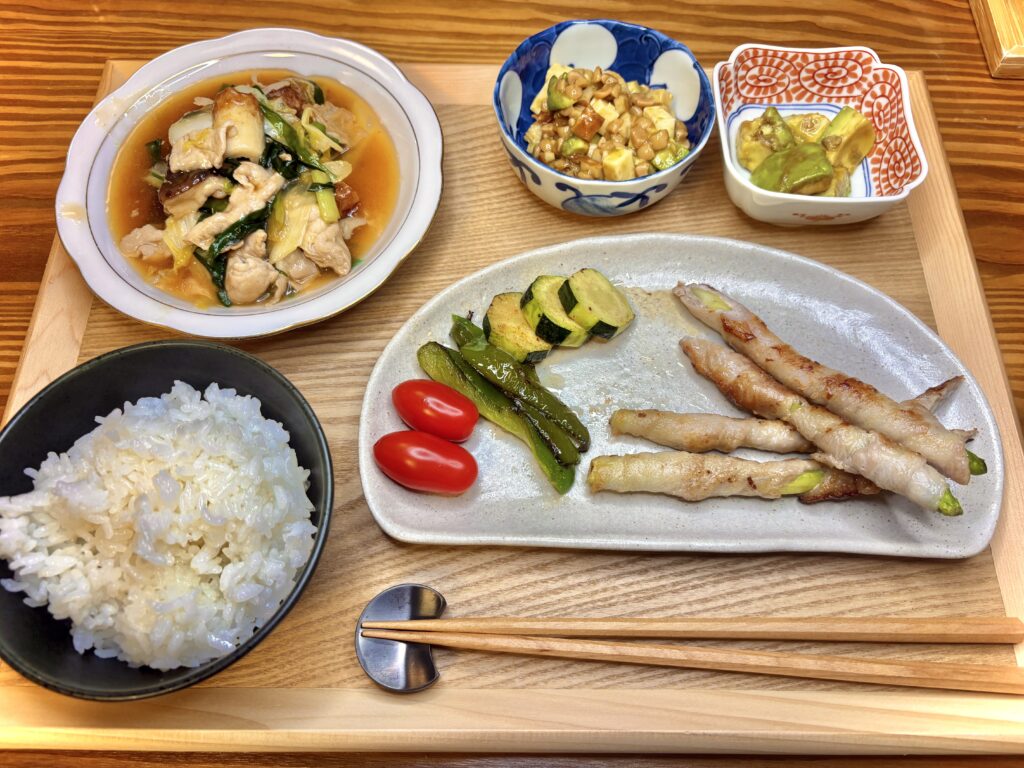 アスパラの豚肉巻きとズッキーニ、ピーマンのグリル、副菜3品とご飯を盛り付けた旦那キッチンの晩ごはん定食。木製トレーに彩りよく並んだ家庭の温かみある食卓。