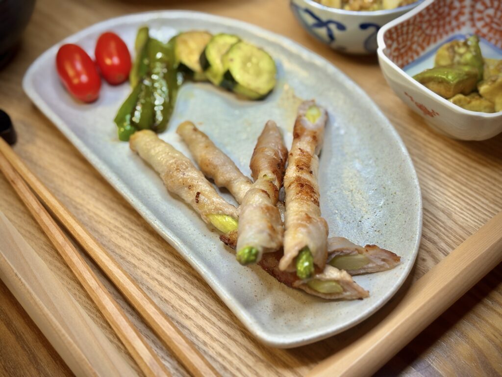 こんがり焼いたアスパラの豚肉巻きを中心に、ズッキーニとピーマンのグリル、ミニトマトを添えた彩り豊かな一皿。木のトレーに並ぶ、旦那キッチンの手作りおかず。