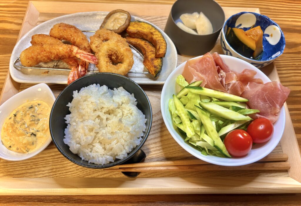 「エビフライとイカリング、カボチャと椎茸のグリル、生ハムサラダ、かぼちゃの煮物、梨を添えた夕食プレート。木のトレーに盛りつけられた彩り豊かな家庭ごはん。