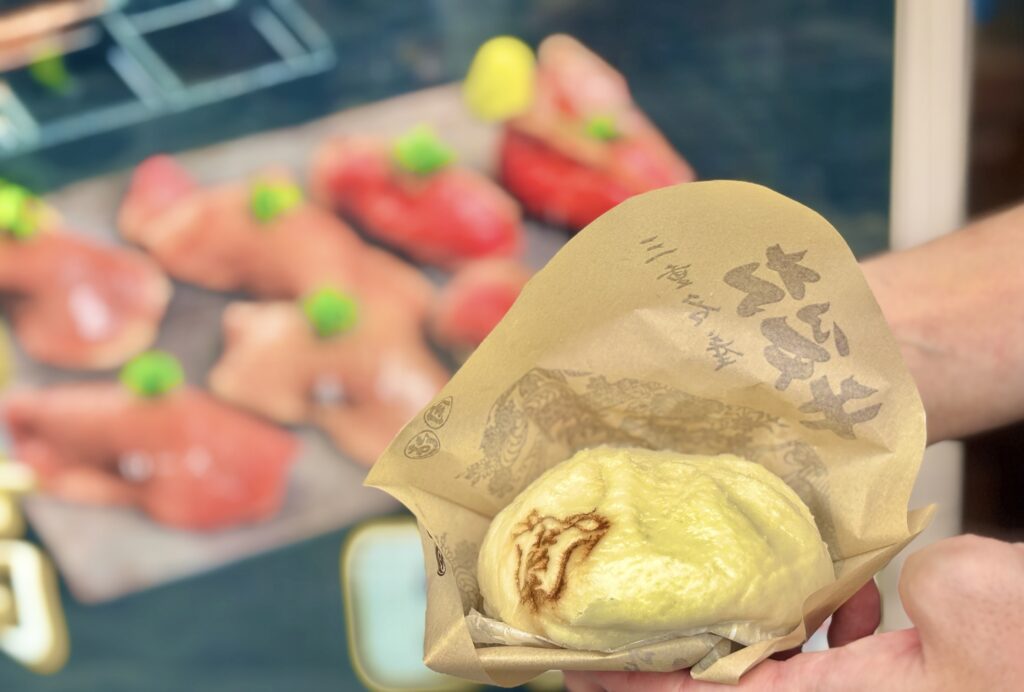 おかげ横丁で販売されている松阪牛まんを手に持つ様子。ふわふわの生地に焼き印が入り、背景には鮮やかな赤身の寿司サンプルが並ぶ。食欲をそそるグルメな一枚。