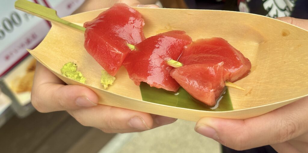 伊勢おかげ横丁で販売されているマグロの串刺し刺身。竹舟型の器に赤々とした新鮮なマグロが並び、わさびが添えられている。手に持って食べ歩きを楽しむ様子。