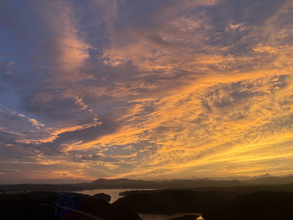 ホテルの部屋から見た伊勢の夕景。雲が黄金色に染まり、空一面に広がるグラデーションが美しい。静かな海と山のシルエットが、まるで絵画のように浮かび上がる幻想的な瞬間。