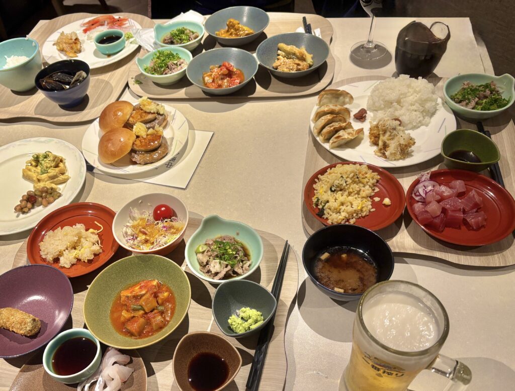 ホテルディナーのビュッフェテーブルに並ぶ多彩な料理。ステーキバーガー、餃子、刺身、サラダ、デザートまで並び、家族でにぎやかに楽しむ夕食