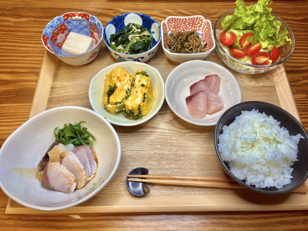 「鰤しゃぶとお造りを中心にした和定食の夕食。青さ入り卵焼き、大根葉としらすのごま油炒め、ほうれん草とえのきのナムル、冷奴、サラダ、小鉢が並ぶ。木のトレイに白ごはんと共に丁寧に盛り付けられた家庭料理の食卓。」