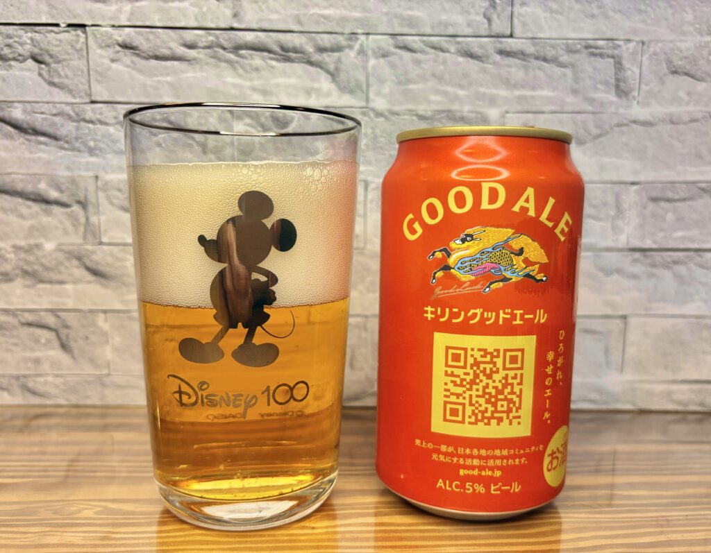 キリンのグッドエールと、ミッキーのシルエットが描かれたDisney100グラスに注がれたビール。黄金色の液体とクリーミーな泡が美しく、明るい木目のテーブルに映える一枚。