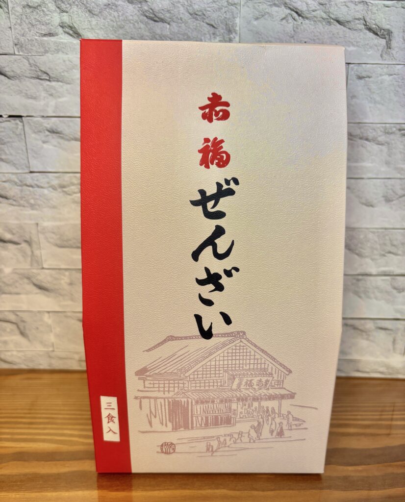 赤福ぜんざいの箱パッケージを撮影した写真。
白地に赤い帯が入った上品なデザインで、「赤福ぜんざい」と大きく書かれている。
下部には赤福本店の建物が描かれ、「三食入」と表示されている。