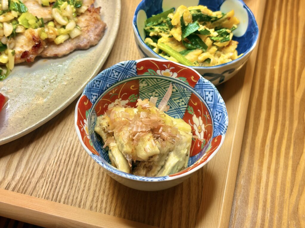 和柄の器に盛られた焼きなす。表面は香ばしく焼かれ、上にはかつおぶしがたっぷり。奥にはニラ玉とポークソテーが並ぶ秋の食卓の一品。