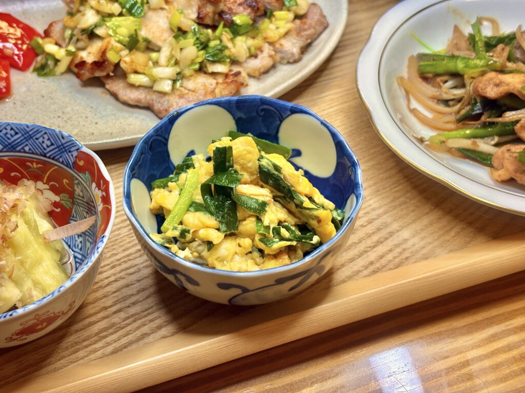 彩り鮮やかなニラ玉。ふわふわの卵にシャキッとしたニラがからみ、和柄の器に盛りつけられている。奥にはポークソテーと焼きなす、亀山ホルモン炒めが並ぶ。