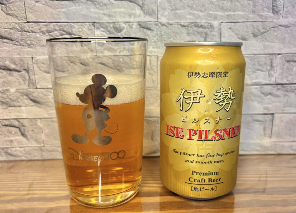伊勢志摩限定「伊勢ピルスナー」の缶と、注がれたビールのグラスを並べた写真。
グラスにはミッキーマウスのシルエットと「Disney100」のロゴが描かれており、
黄金色のビールが美しく透けている。