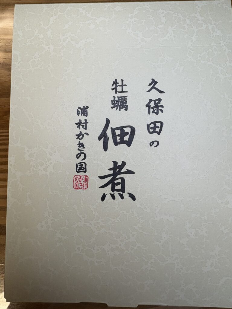 久保田の牡蠣佃煮」の化粧箱。
上品な和紙風の白いパッケージに、黒い筆文字で「久保田の牡蠣佃煮」「浦村かきの国」と書かれている。
中央に大きく「佃煮」の文字があり、落款の赤印が添えられている。