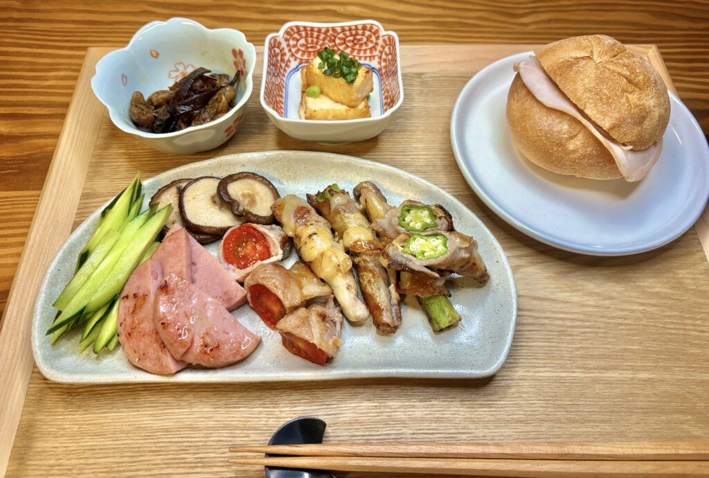 エノキとオクラの豚肉巻き、ミニトマトの豚肉巻き、ボロニアソーセージ、しいたけソテー、厚揚げ焼き、伊勢みやげの牡蠣の佃煮、ダンマルシェのカイザーパンが並ぶ和洋折衷プレートの夕食
