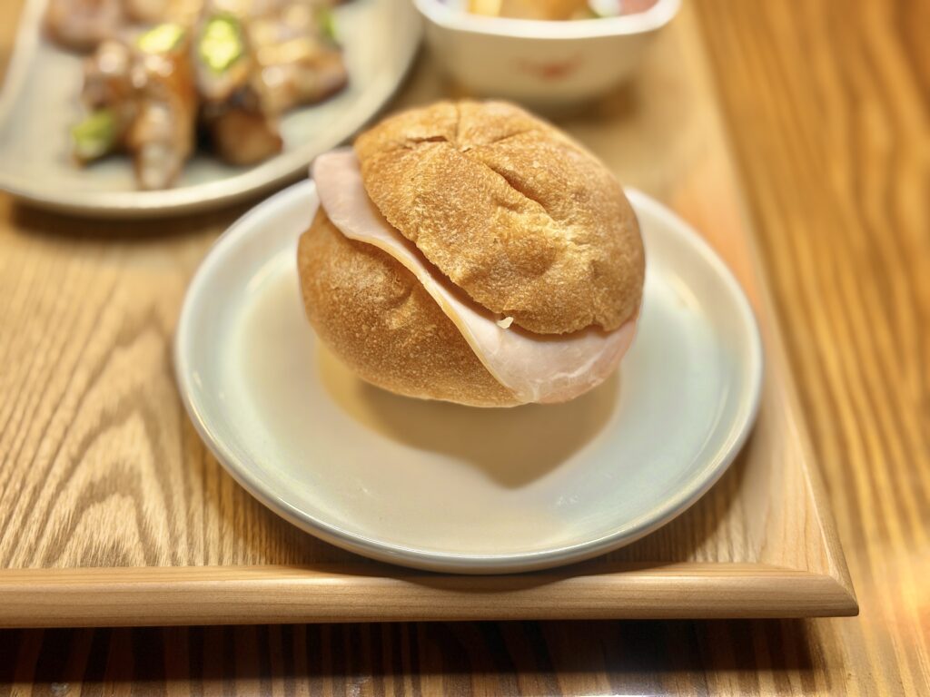 ダンマルシェのカイザーパンにハムを挟んだシンプルなサンドが白い皿にのせられた様子。奥には豚肉巻きや厚揚げ焼きが並ぶ木のトレーがぼんやりと映る