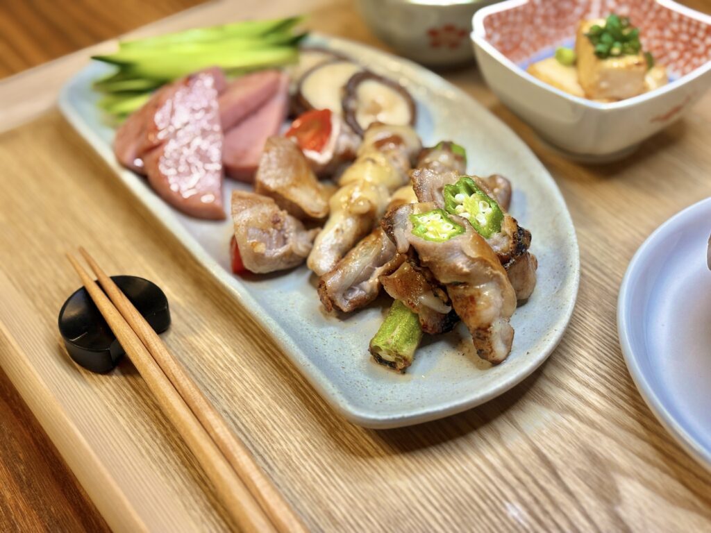 オクラとエノキの豚肉巻き、ミニトマトの豚肉巻き、ボロニアソーセージ、しいたけソテーなどが美しく盛り付けられたプレート。木のトレーに箸が添えられた和洋ミックスの夕食