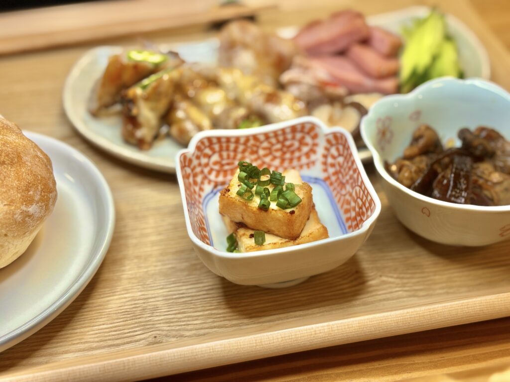 厚揚げ焼きに刻みねぎをのせた小鉢を中心に、隣に伊勢みやげの牡蠣の佃煮、奥には豚肉巻きやボロニアソーセージ、きゅうりスティックが並ぶプレート。手前にダンマルシェのカイザーパンが添えられた温かみのある夕食シーン