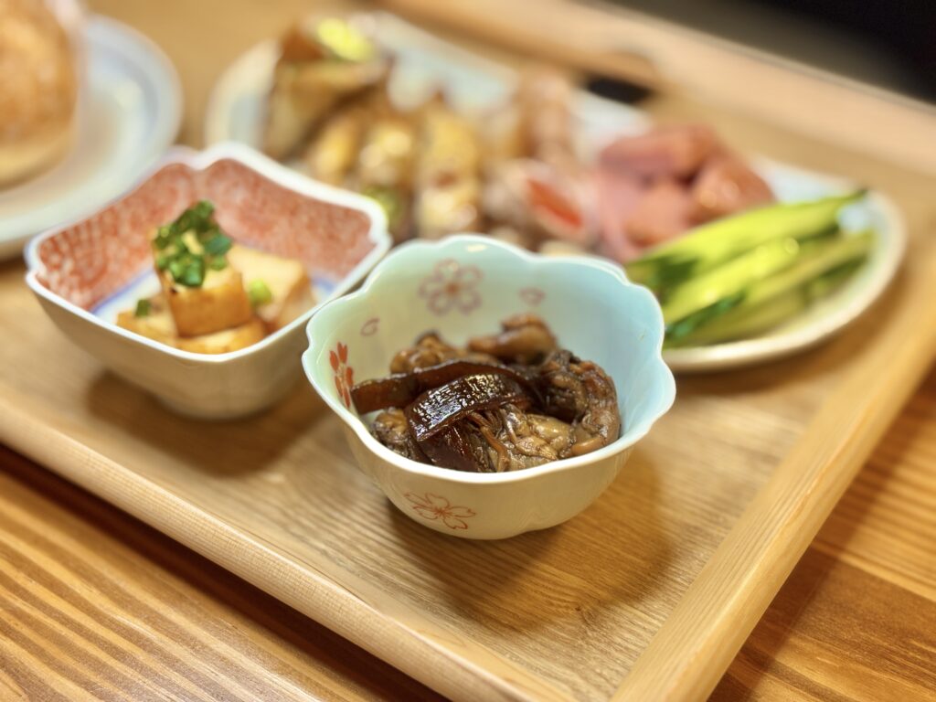 伊勢みやげの牡蠣の佃煮と厚揚げ焼きが小鉢に盛られ、奥には豚肉巻きやボロニアソーセージ、きゅうりスティックが並ぶプレート。木のトレーに温かみのある照明が映える和風の食卓