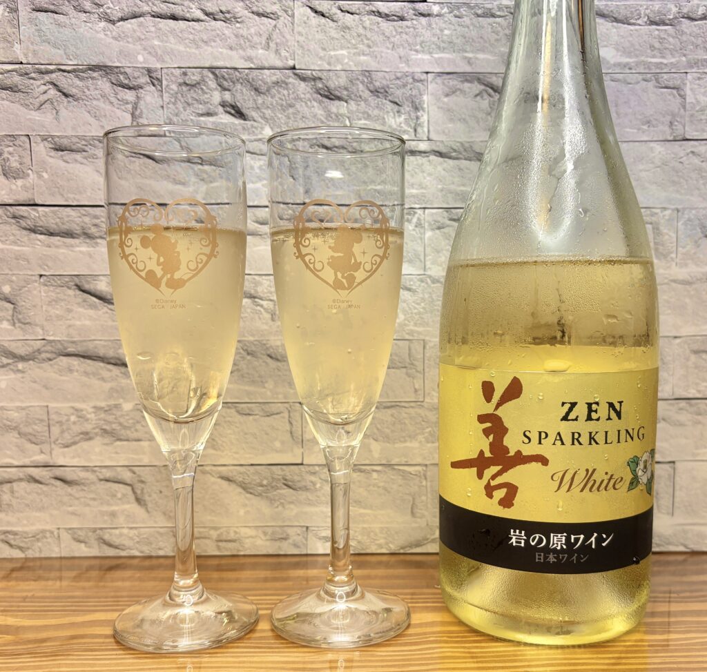 岩の原ワイン『善 ZEN スパークリング ホワイト』のボトルと、泡立つスパークリングワインが注がれた2つのシャンパングラス。木のテーブルと石目調の背景が上品な雰囲気を演出している