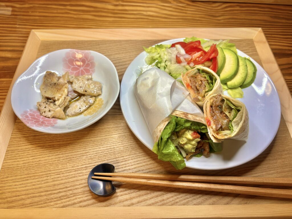 木製トレイの上に盛り付けられた夕食。右の白いプレートには、プルコギアボカドタコスが2本と、アボカド・トマト・レタスのサラダが並んでいる。左の花柄の小鉢には、伍魚福の鯛のバジルオリーブオイル漬け。全体が温かみのある照明に包まれた、おうちカフェ風の食卓。