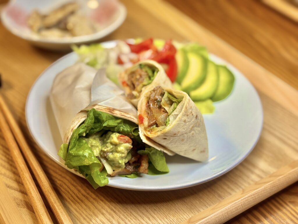 白いプレートに盛られたプルコギアボカドタコス。手前のタコスにはたっぷりのアボカドとレタス、奥には甘辛プルコギの具材が見える。横にはスライスしたアボカドとトマト、レタスが彩りよく添えられ、奥に鯛のバジルオリーブオイル漬けの小鉢がぼんやり映り込んでいる。温かみのある照明に照らされた、おうちレストラン風の一皿。