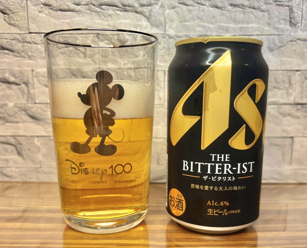 ミッキー柄のグラスに注がれた黄金色のビールと、黒×金デザインの「ASAHI THE BITTER-IST」缶。泡立ちが美しく、上品な印象の一枚。