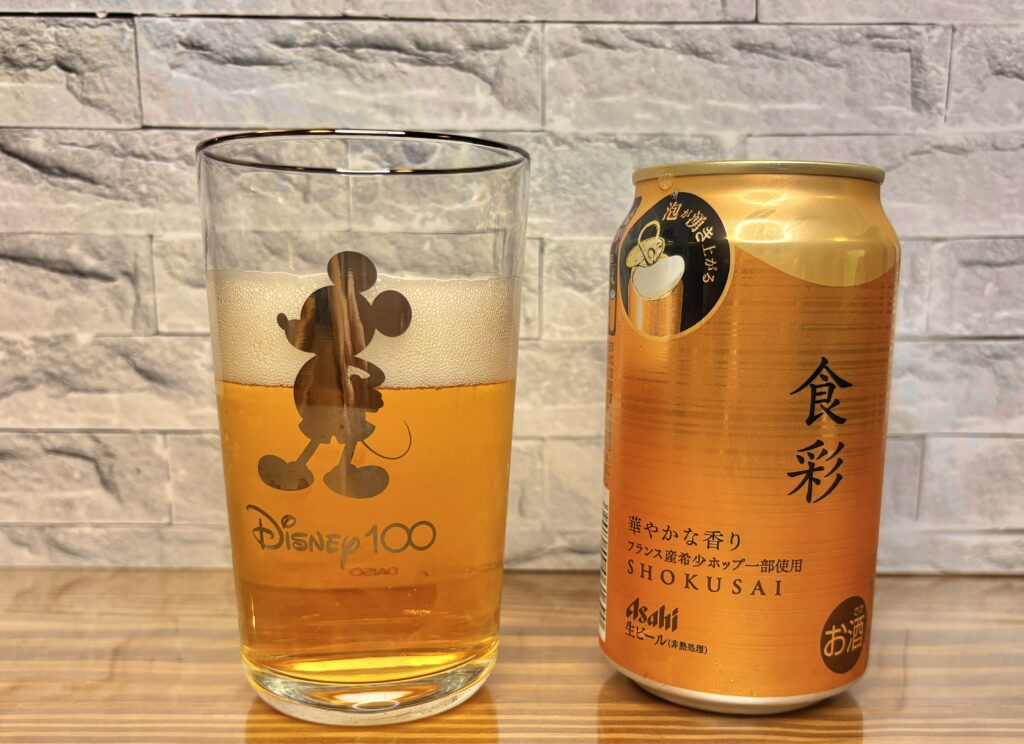 アサヒ生ビール『食彩』の缶と、注がれたビールが入ったミッキーのグラス。黄金色のビールが泡立ち、テーブルに置かれている。