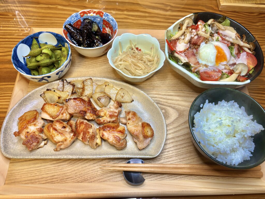 阿波尾鶏のグリルを中心に、温玉シーザーサラダ、黒枝豆、ナスの煮浸し、もやしのナムル、白ごはんが並ぶ晩ごはん。木目のトレーの上に、彩り豊かな家庭料理が整然と並んでいる