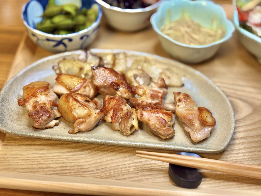 香ばしく焼き上げた阿波尾鶏のグリル。皮はパリッと黄金色に焼け、玉ねぎのソテーと一緒に盛り付けられた一皿。背景には黒枝豆やナスの煮浸しなど副菜も並ぶ。