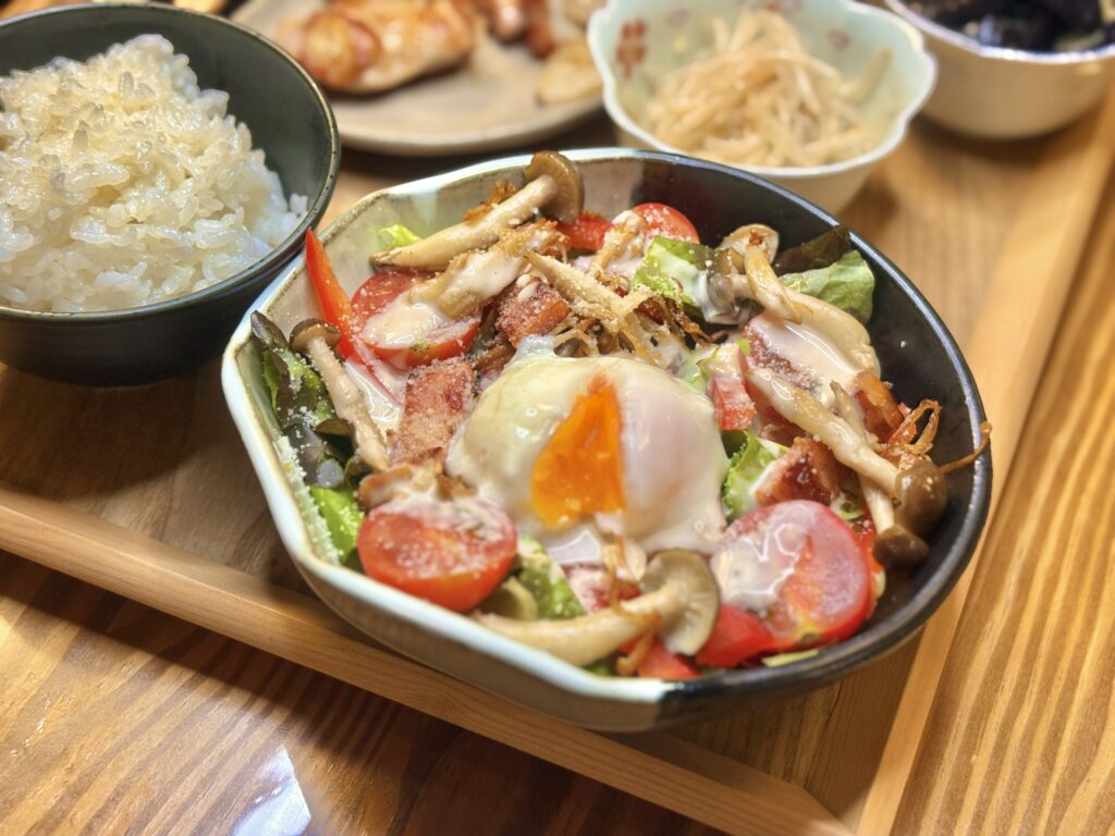 温泉卵をのせたシーザーサラダ。ベーコン、しめじ、トマト、レタスが彩りよく盛り付けられ、卵の黄身がとろりと流れ出す直前の瞬間。隣には白ごはんと副菜が並ぶ。